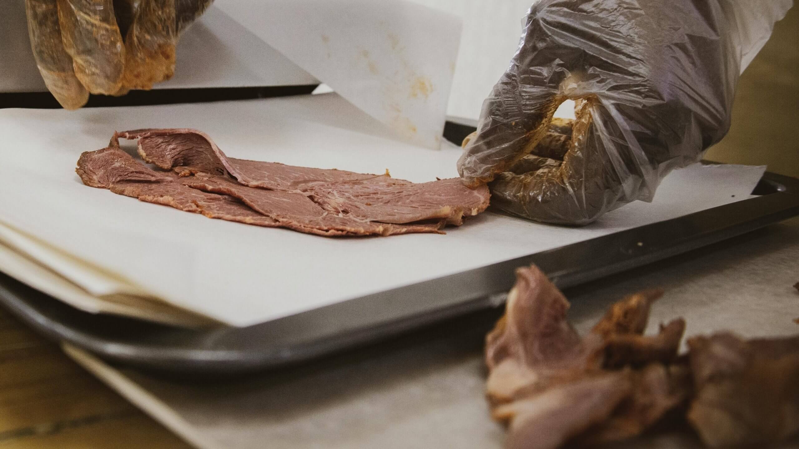 Gloved hands slicing thin pieces of cooked meat on parchment paper over a metal tray.