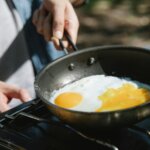 Person frying two eggs in a black pan on a gas stove outdoors