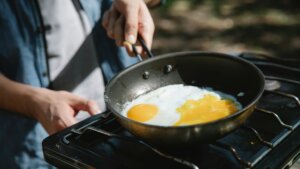 Person frying two eggs in a black pan on a gas stove outdoors