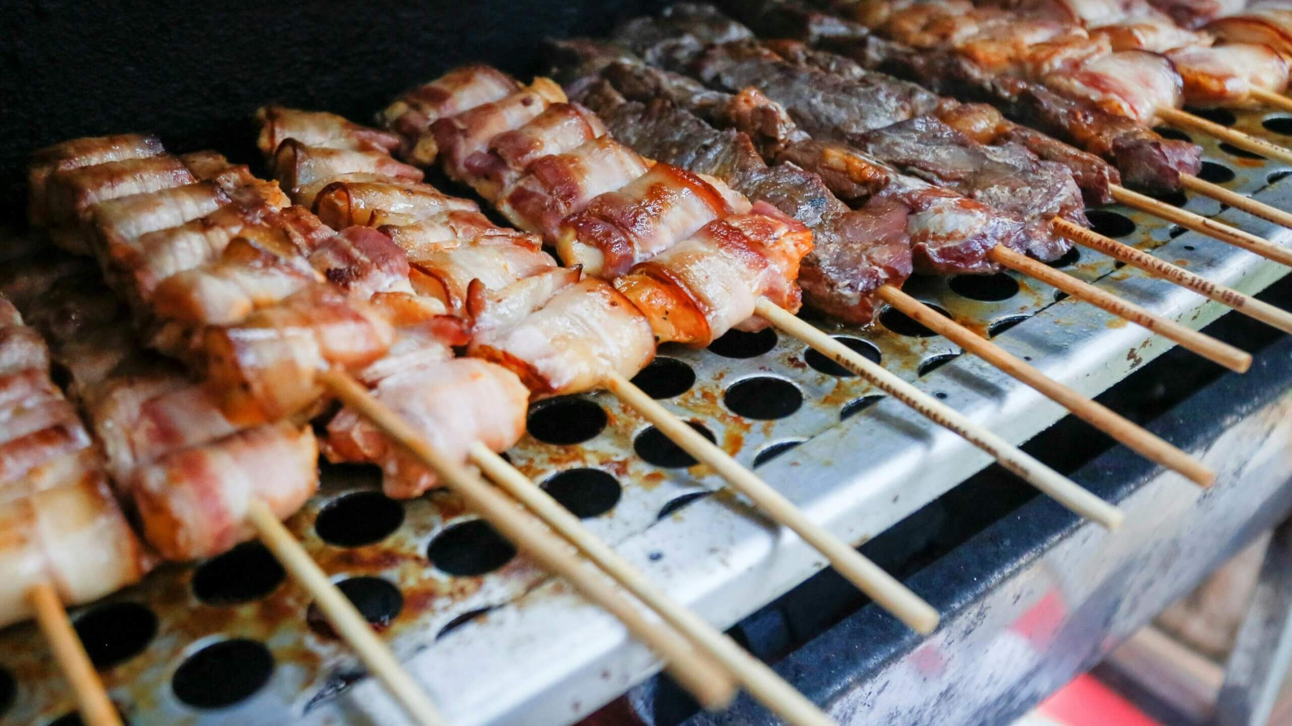 Grilled bacon-wrapped and beef skewers cooking on a metal barbecue grill rack
