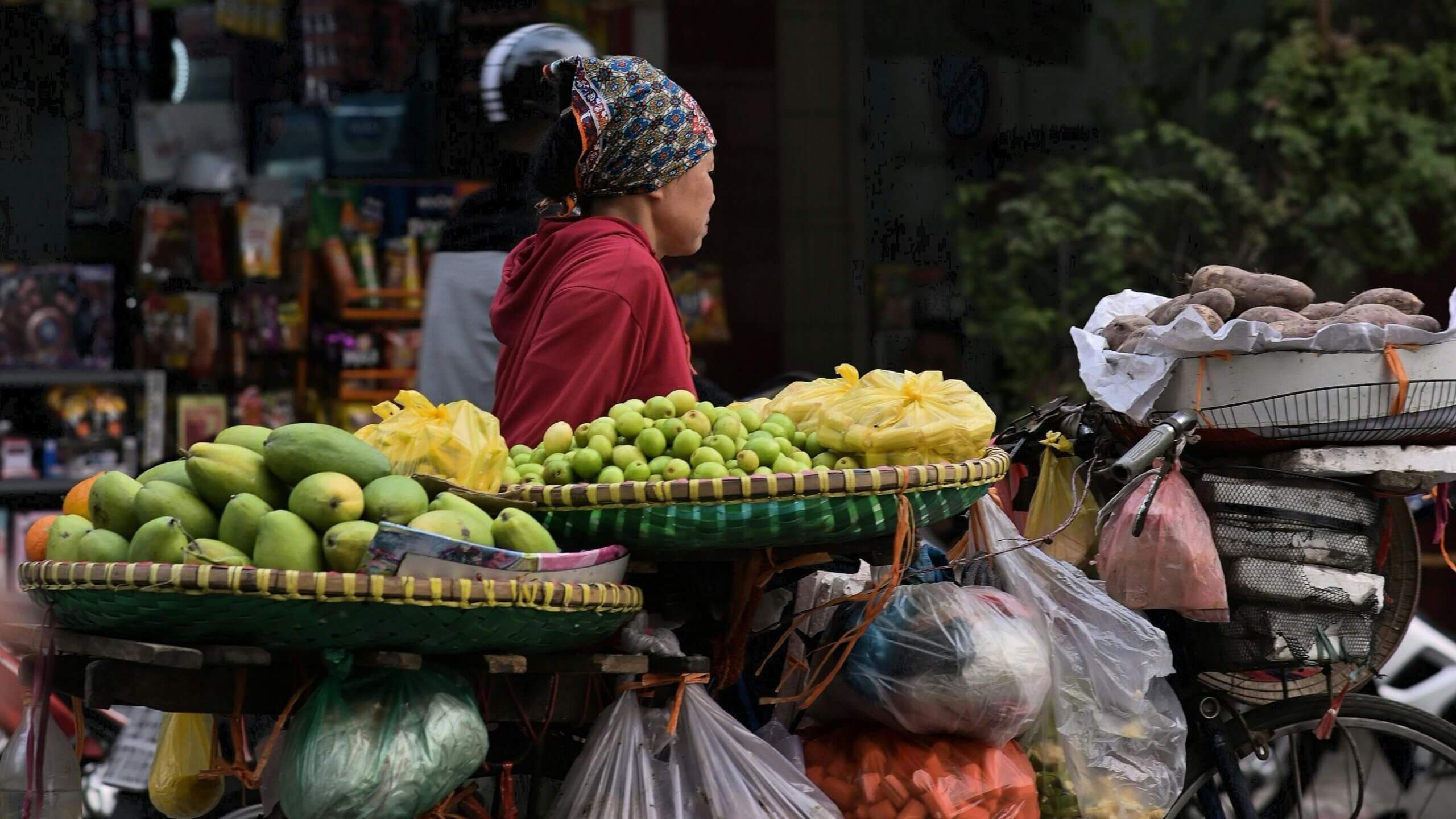 Woman wearing a patterned headscarf and red hoodie selling mangoes and other produce from a bicycle cart.