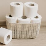 Six white toilet paper rolls, five stacked in a beige woven basket and one lying on a wooden floor.