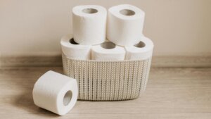Six white toilet paper rolls, five stacked in a beige woven basket and one lying on a wooden floor.