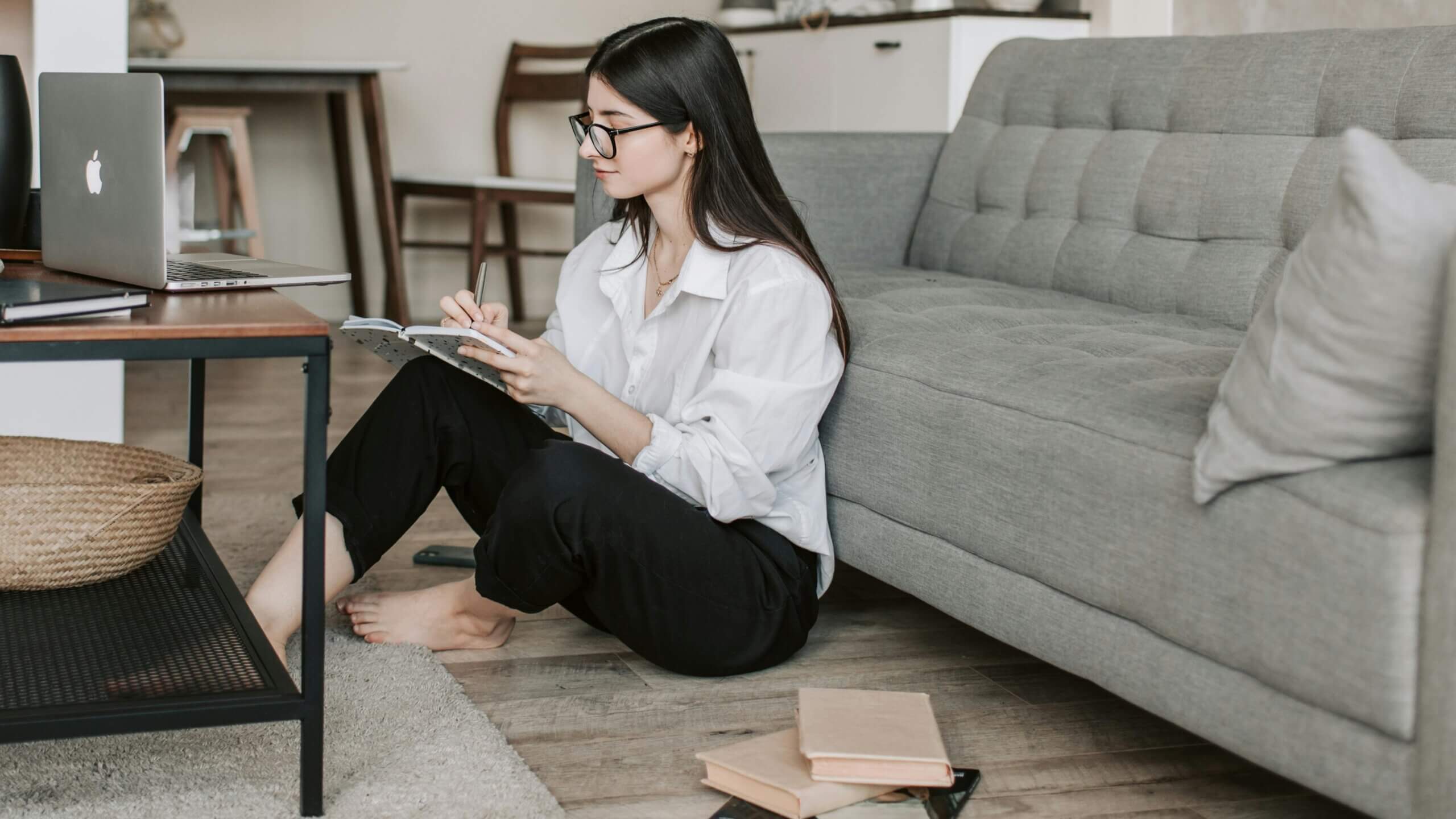Young woman with glasses sitting on the floor writing in a notebook next to a MacBook on a coffee table