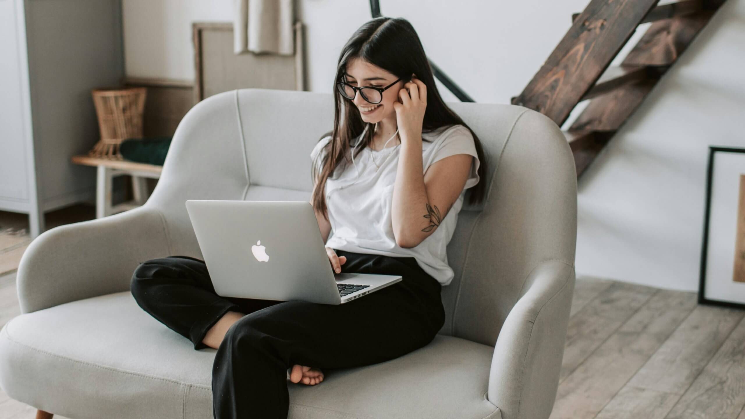 Young woman with glasses sitting cross-legged on a gray couch using a silver Apple MacBook laptop and wearing earphones