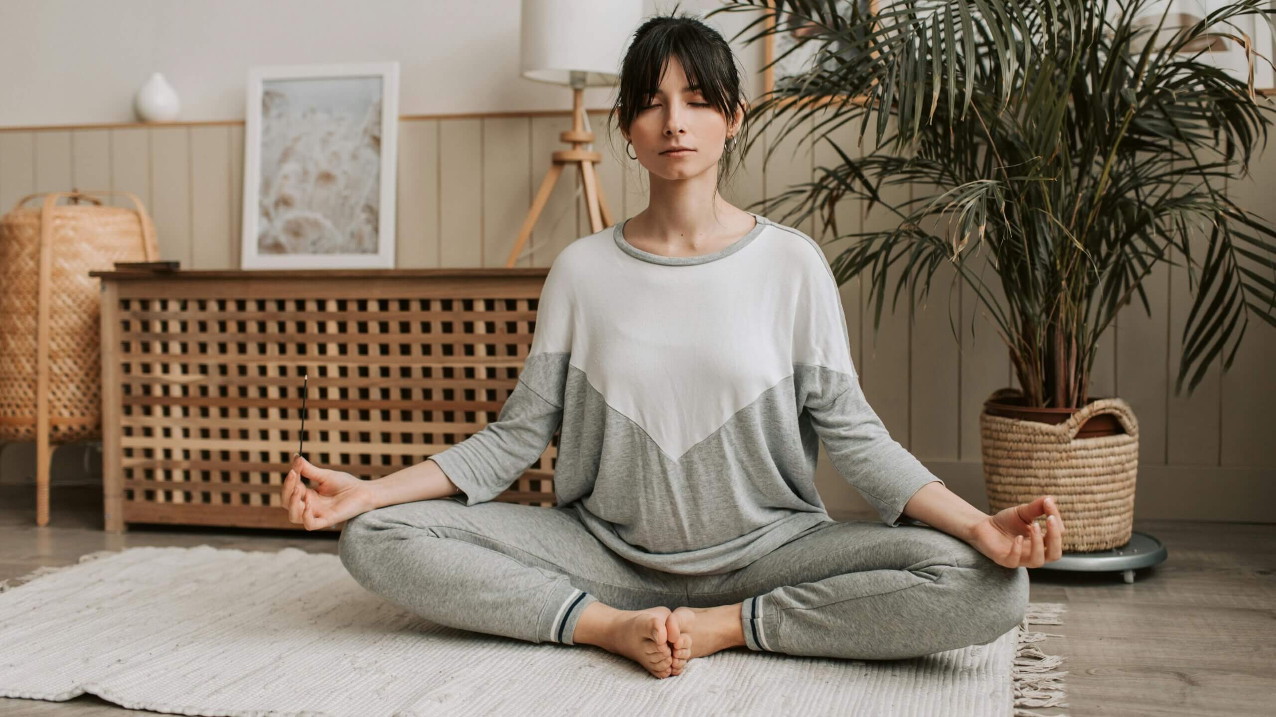 Woman in gray and white loungewear meditating cross-legged on a rug in a cozy room with plants and wooden furniture