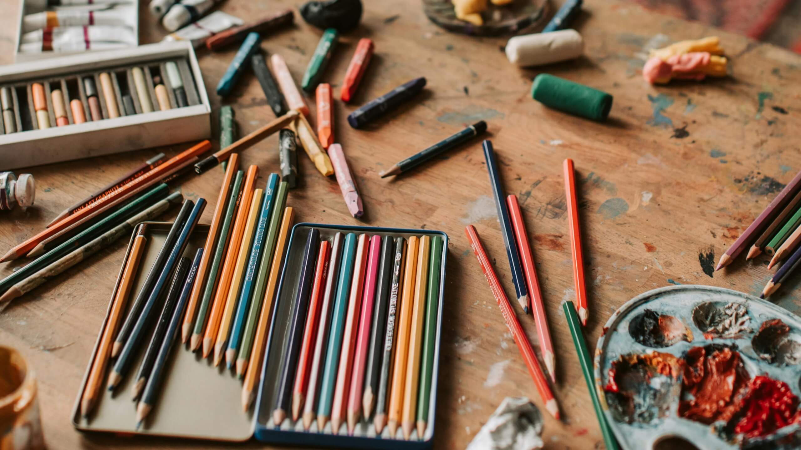 Colored pencils, pastels, and a paint palette scattered on a wooden artist's table