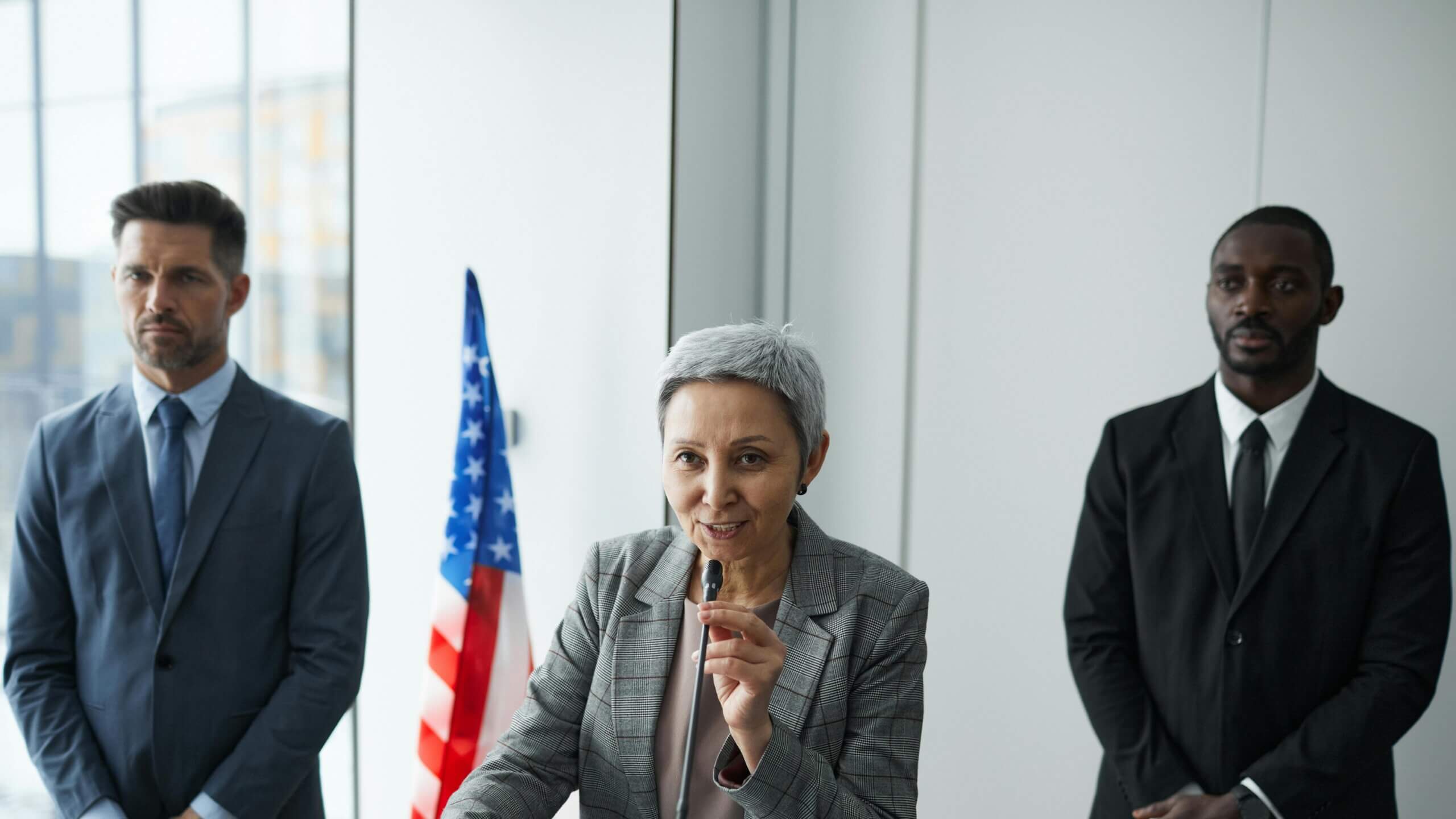 Woman speaking at a podium with a microphone, flanked by two men in suits and an American flag in the background.