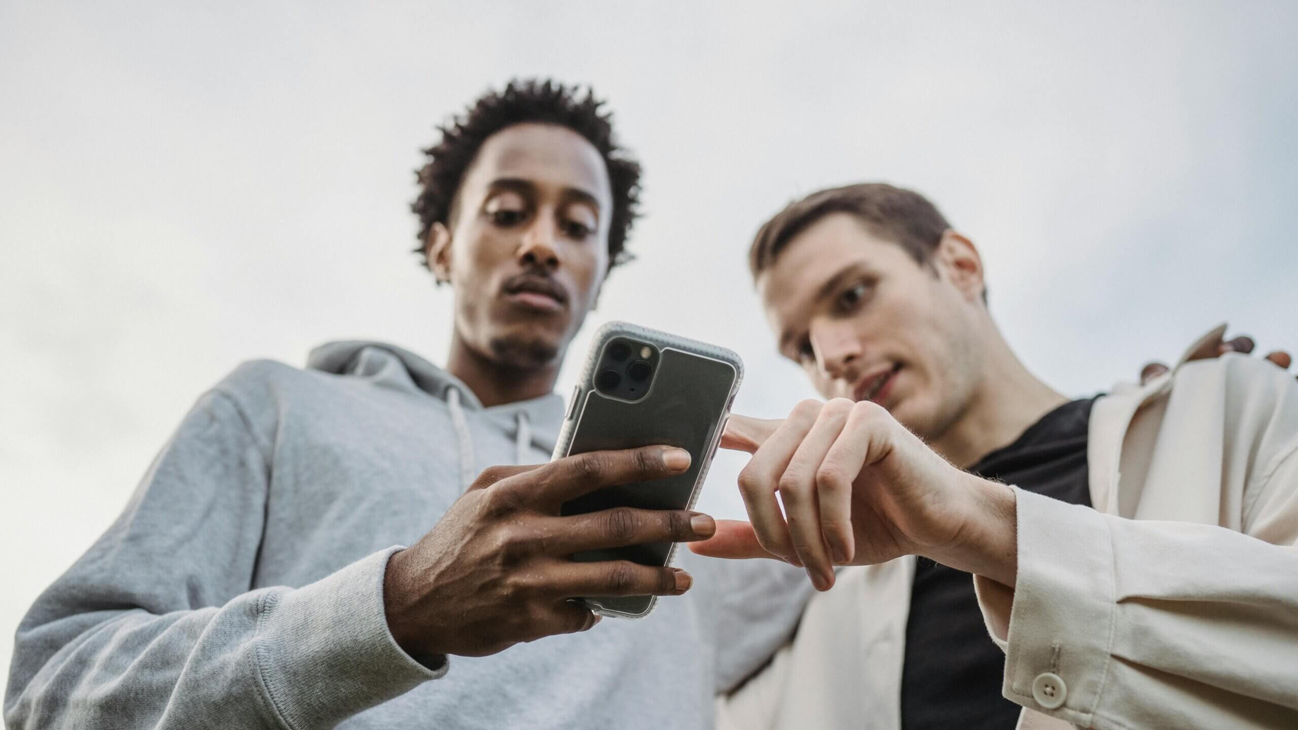 Two men looking at and interacting with a smartphone outdoors against a cloudy sky.