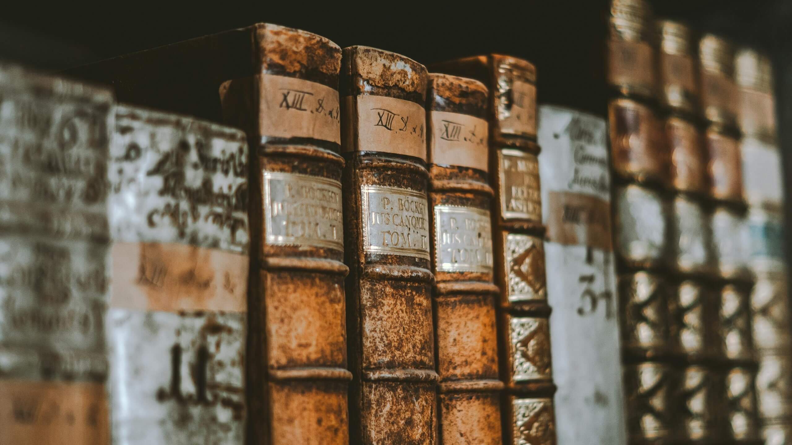 Close-up of old leather-bound books with Latin titles on a wooden shelf.