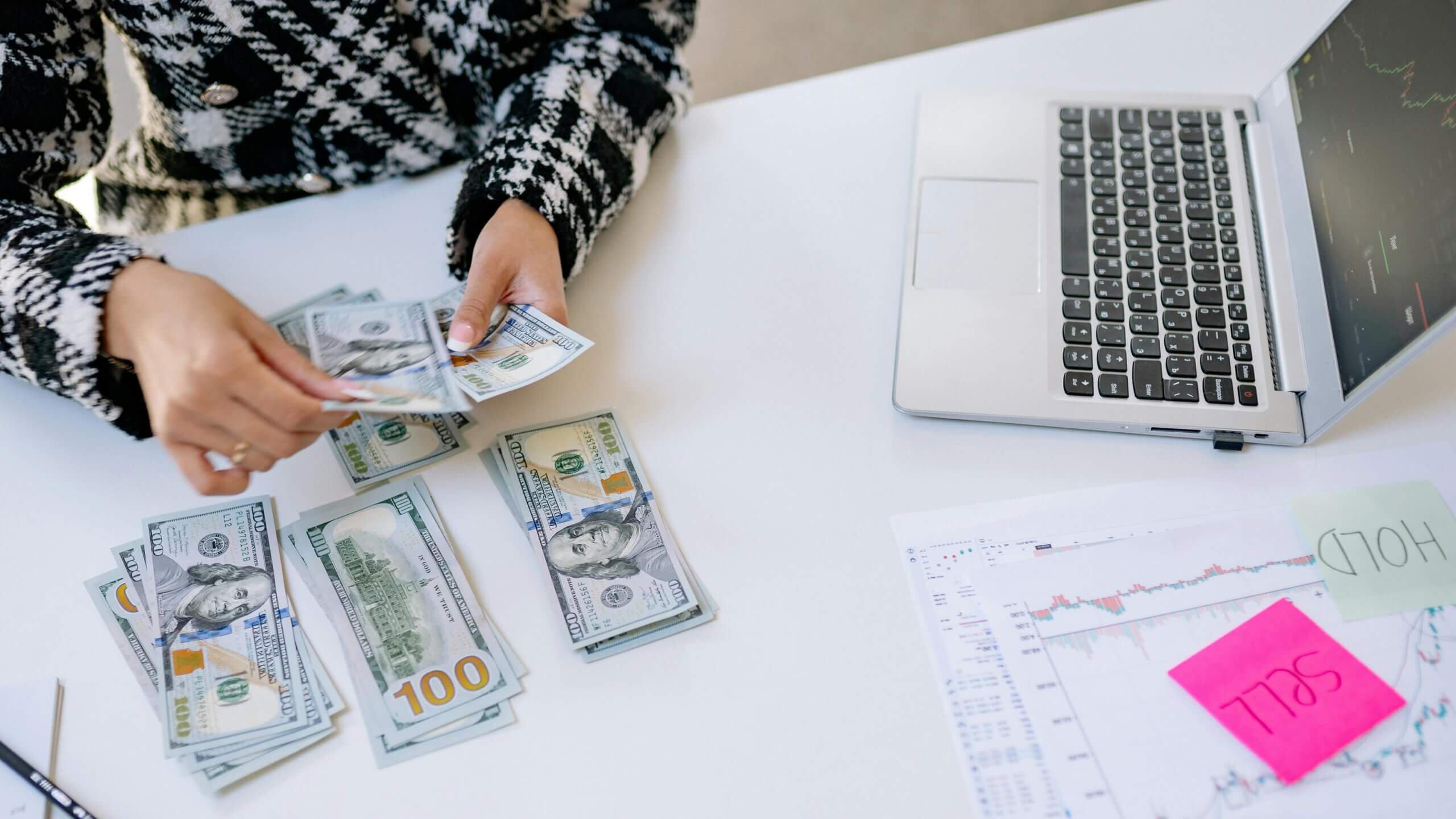 Person counting US hundred-dollar bills next to a laptop displaying stock charts and papers with "SELL" and "HOLD" notes.