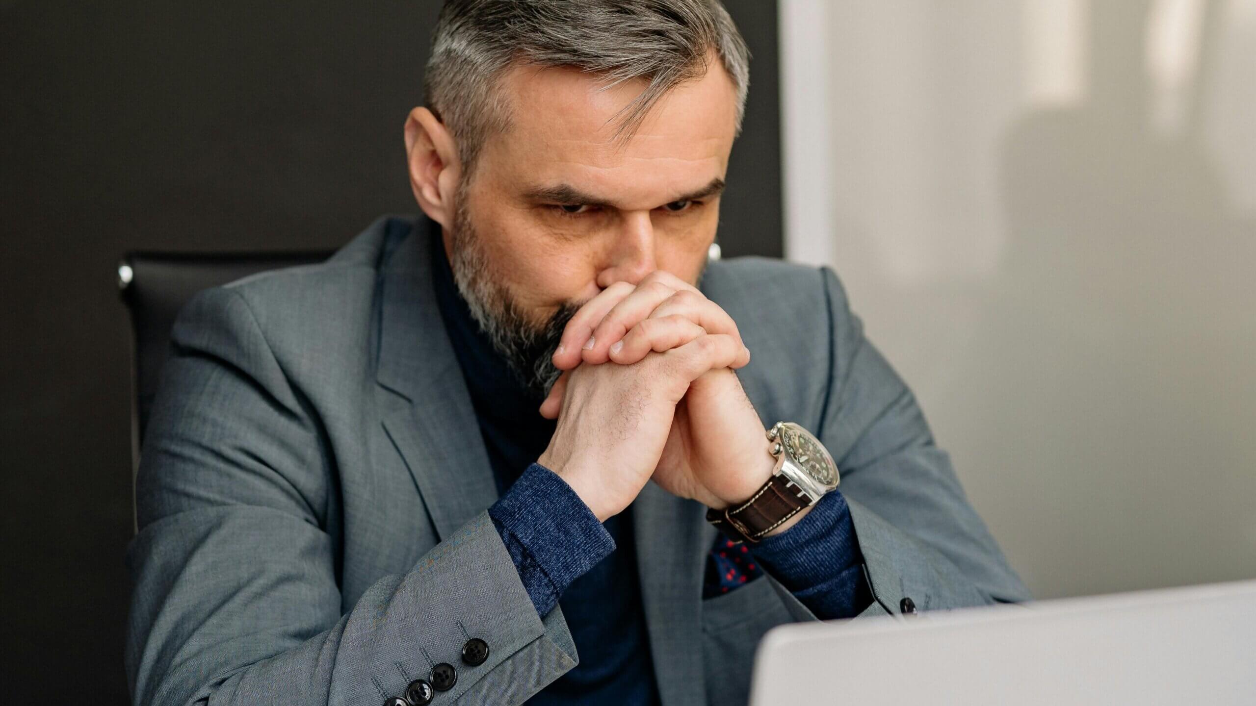 Man in gray suit and blue shirt sitting at desk, looking thoughtful with hands clasped near face