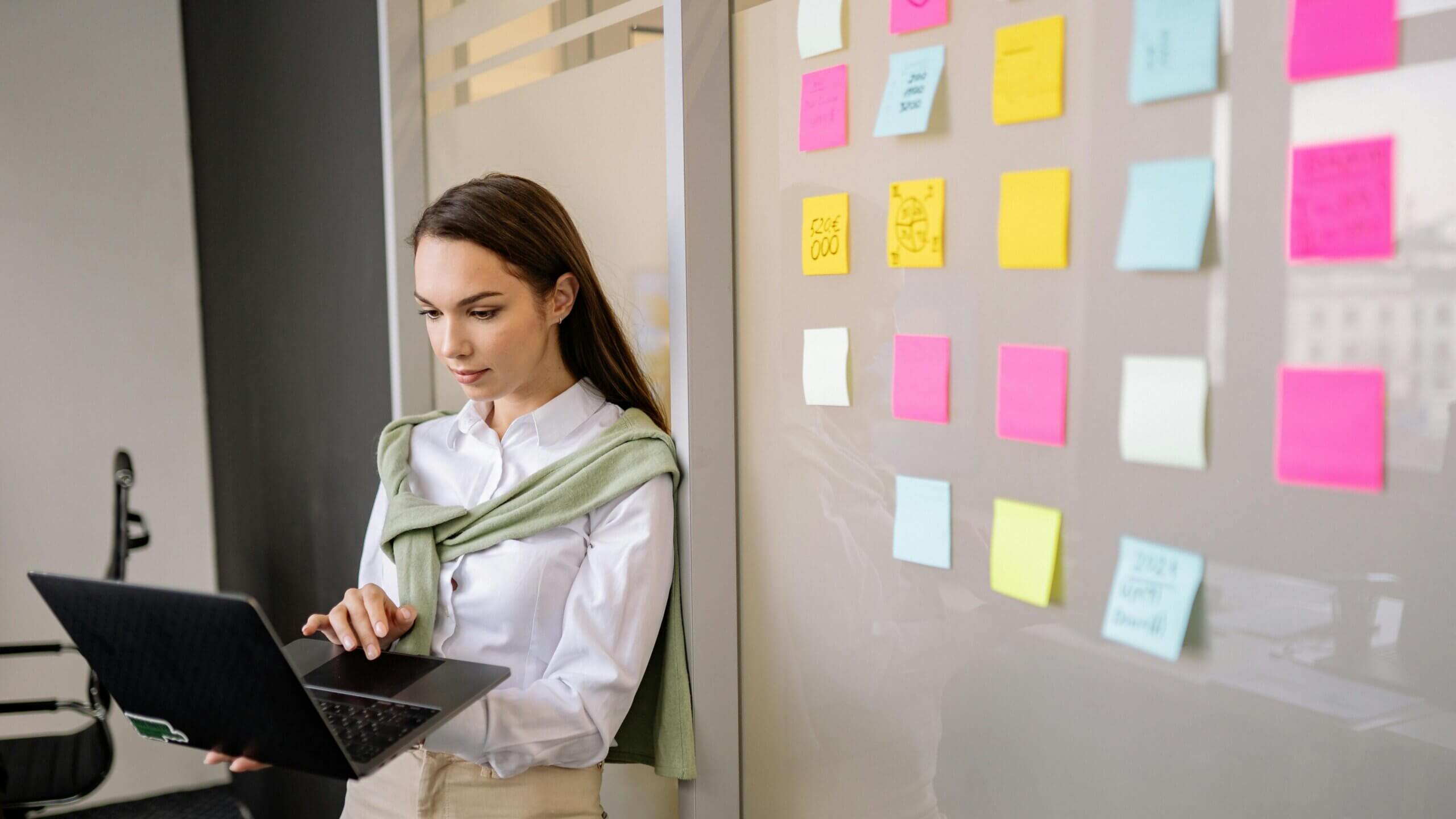 Woman in white shirt and green sweater using laptop near glass wall with colorful sticky notes