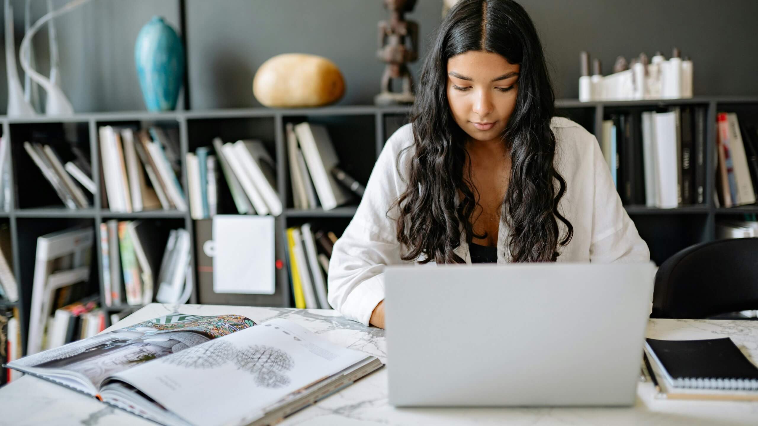 Young woman with long dark hair working on a laptop at a desk with open books and a bookshelf in the background