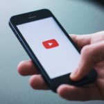 Hand holding a smartphone displaying the YouTube app loading screen with the red play button logo.