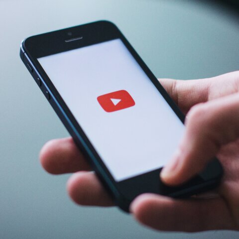 Hand holding a smartphone displaying the YouTube app loading screen with the red play button logo.