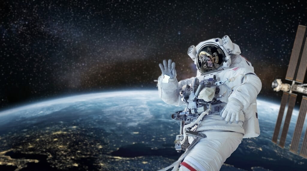 Astronaut in white spacesuit waving during a spacewalk with Earth and the International Space Station in the background