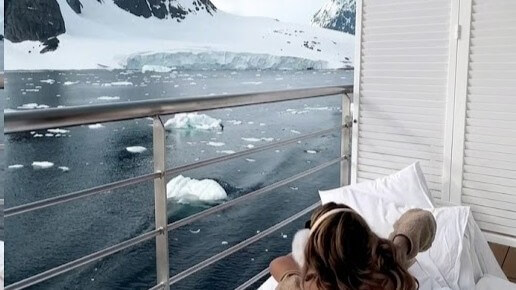 Person reclining on a bed on a balcony overlooking icy water and snow-covered mountains.