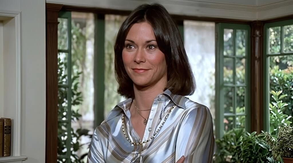 Woman with short brown hair wearing a striped satin blouse standing in a room with large windows and plants outside.