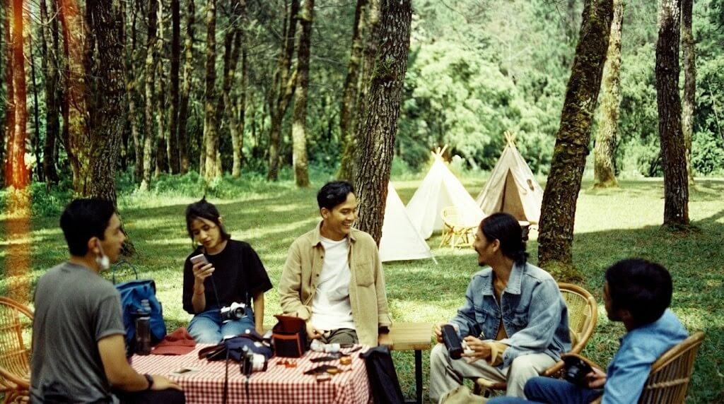 Group of five young adults sitting and chatting around a picnic table in a forest campsite with tents in the background
