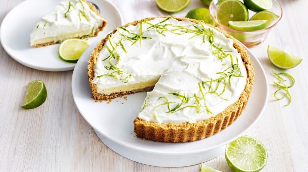 Key lime pie with whipped cream and lime zest, a slice served on a plate with lime wedges around.