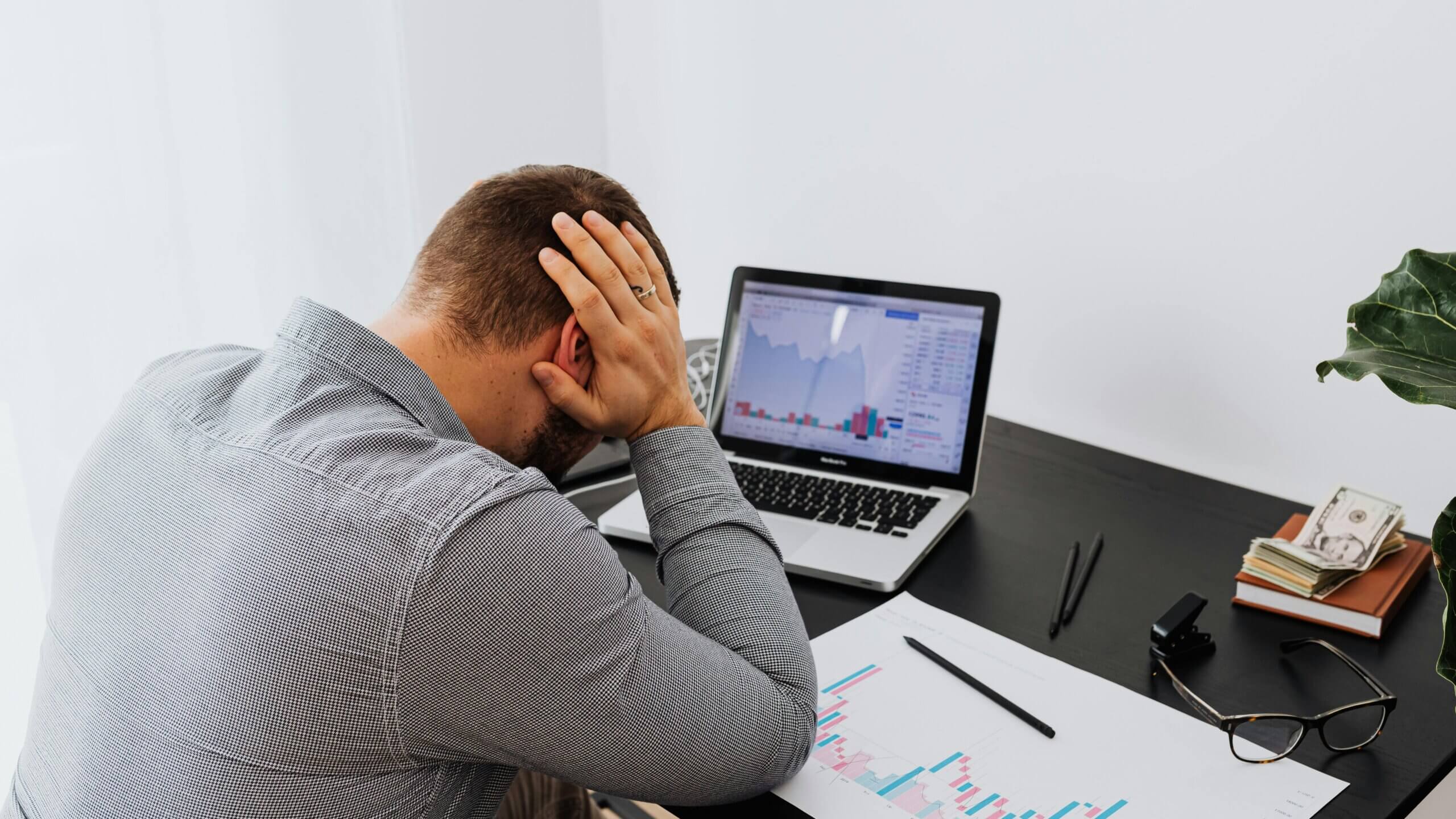 Man in gray shirt holding head at desk with laptop showing stock charts and financial documents