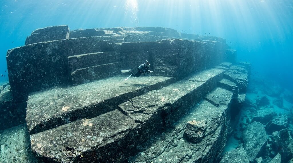 Scuba diver exploring large underwater stone terraces resembling ancient ruins.