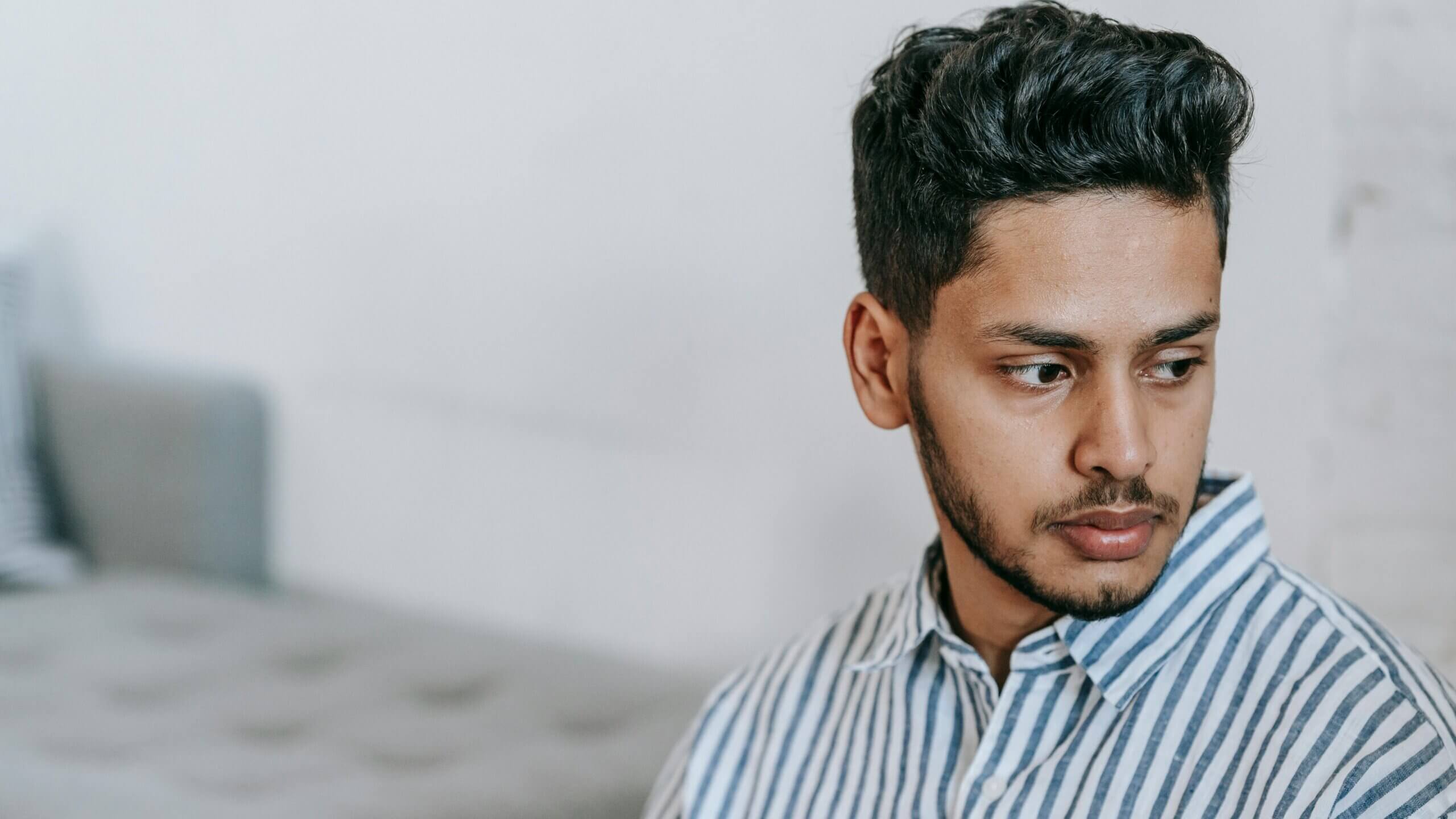 Young man with dark hair and beard wearing a blue and white striped shirt looking to the side indoors
