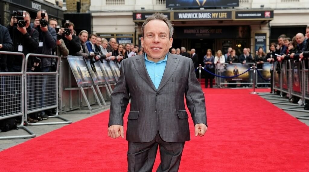 Warwick Davis in a gray suit on a red carpet at the premiere of "Warwick Davis: My Life" with photographers and crowd behind him.
