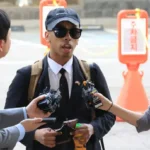 Man wearing sunglasses and a cap being interviewed by reporters holding microphones and recorders outdoors
