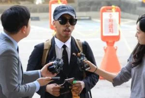 Man wearing sunglasses and a cap being interviewed by reporters holding microphones and recorders outdoors