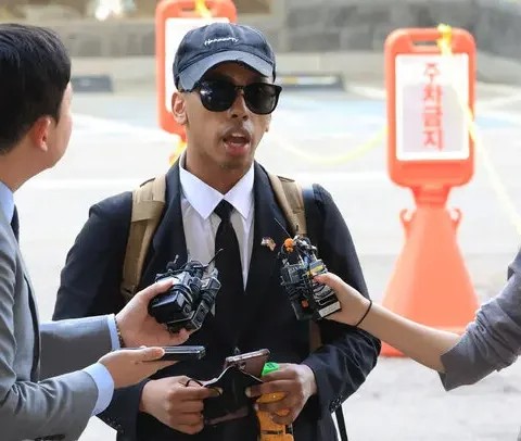 Man wearing sunglasses and a cap being interviewed by reporters holding microphones and recorders outdoors
