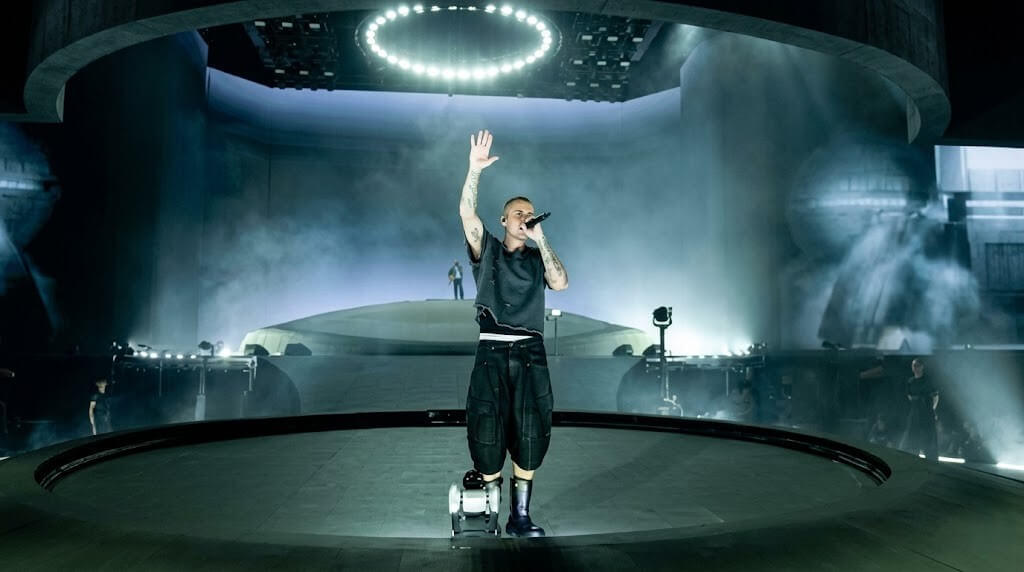 Eminem performing on stage with a microphone under circular stage lights and fog effects