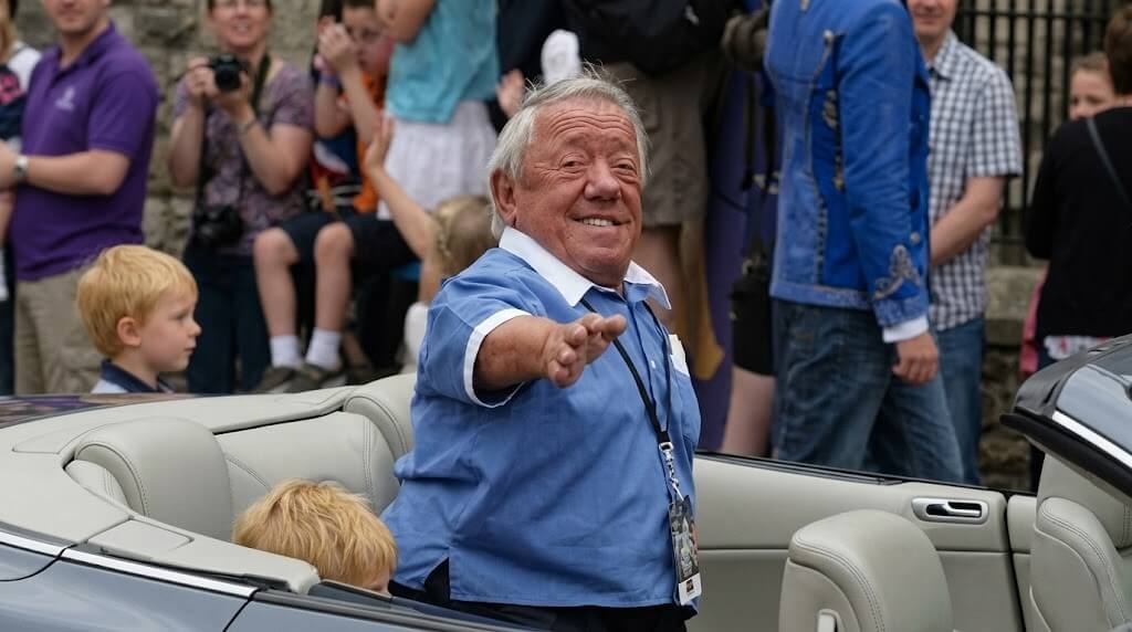 Older man in blue shirt waving from a convertible car during a public event with spectators in the background