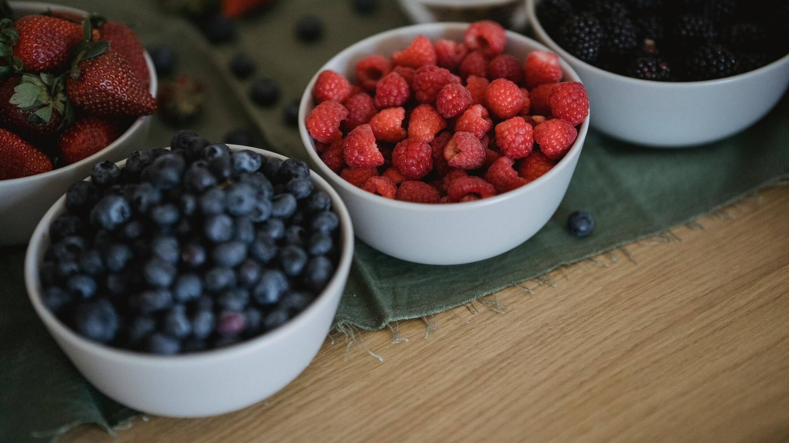 White bowls filled with blueberries, raspberries, strawberries, and blackberries on a green cloth and wooden table.