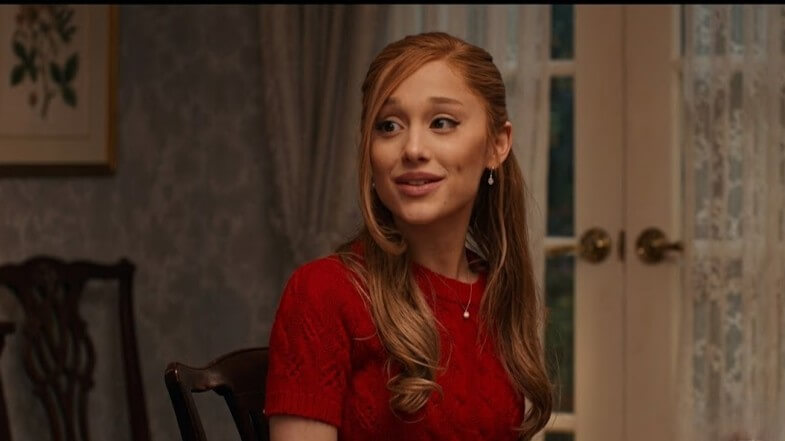 Young woman with long red hair wearing a red sweater sitting indoors near a door with lace curtains