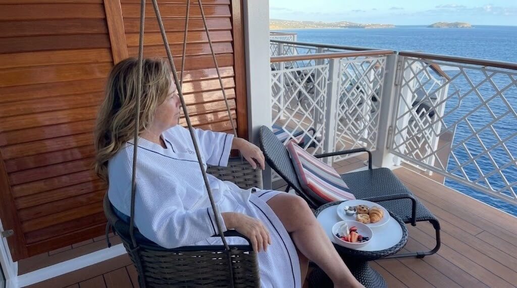 Woman in white robe sitting on a balcony swing overlooking the ocean with a table of pastries and fruit nearby
