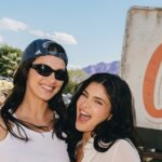 Two smiling women, one wearing a blue Adidas cap and sunglasses, posing outdoors at a sunny event.