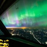 Northern lights visible from an airplane cockpit flying over a city at night.