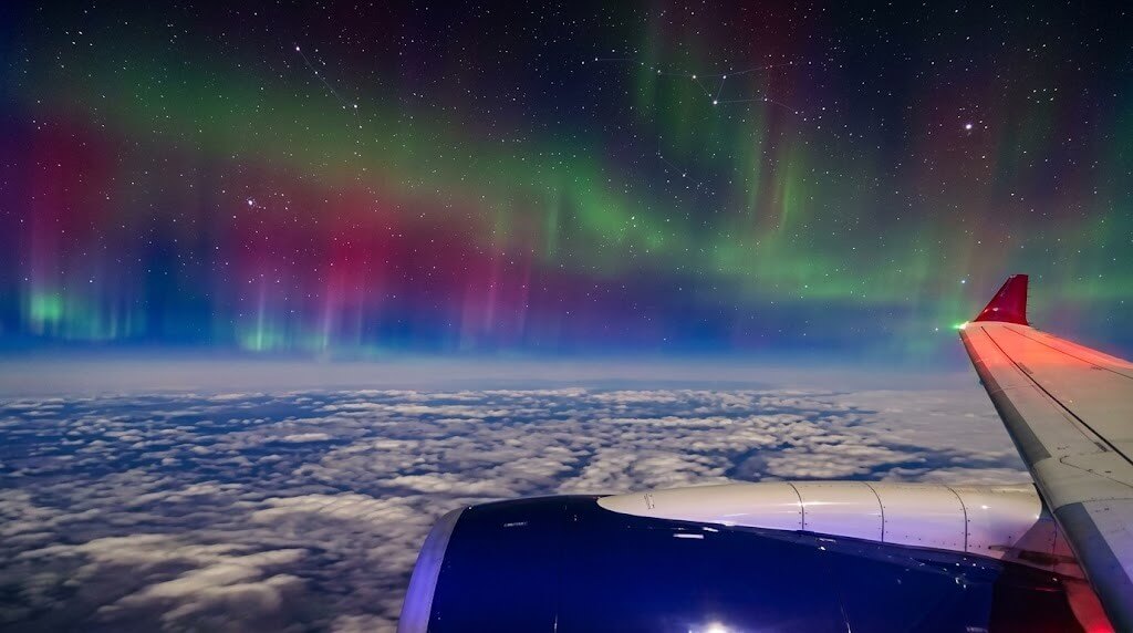 Northern lights visible above clouds from airplane window showing wing and engine at night