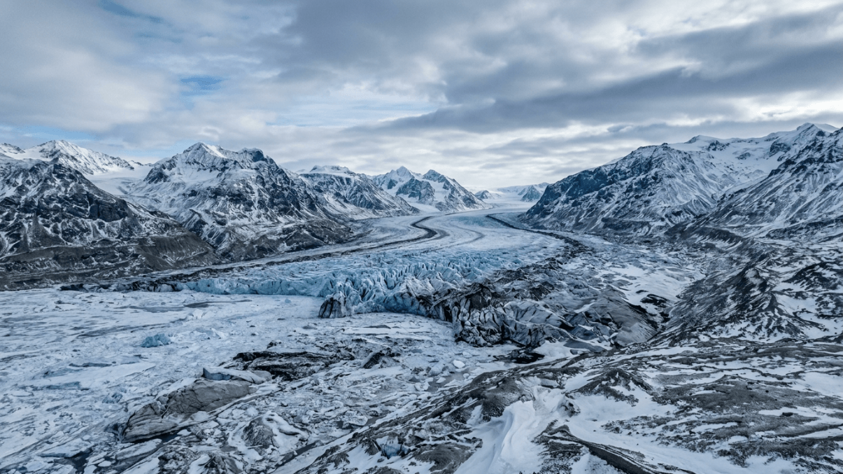 Aerial view of a vast glacier flowing through snow-covered mountains under a cloudy sky.