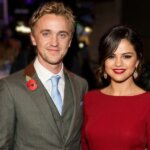 Tom Felton in a gray suit with a poppy pin and Selena Gomez in a red dress at a formal event.