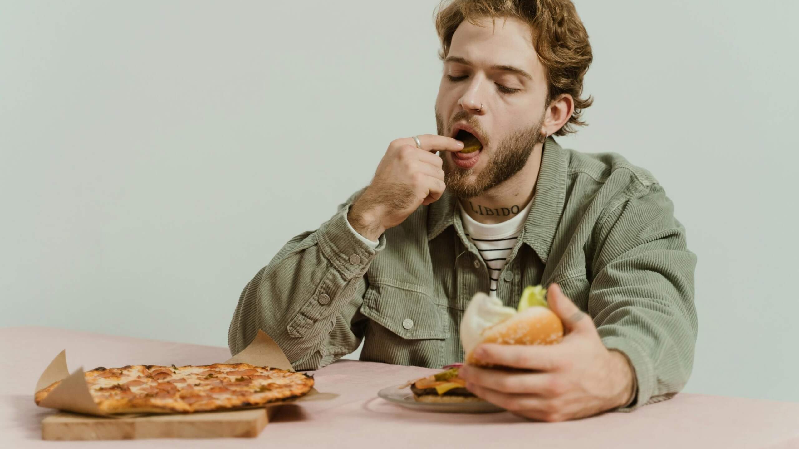 Man with beard and nose ring eating a burger and licking his finger with a pizza on the table in front of him