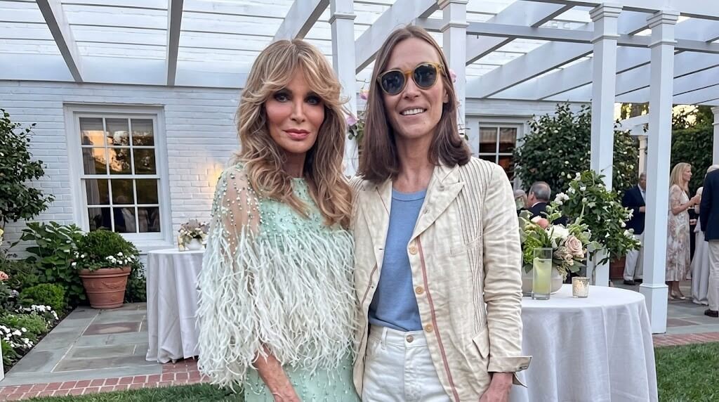 Two women posing outdoors at a garden event, one wearing a light green feathered dress and the other in a beige jacket and sunglasses.