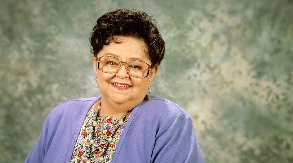 Smiling elderly woman with glasses wearing a purple cardigan and floral blouse against a mottled green background.