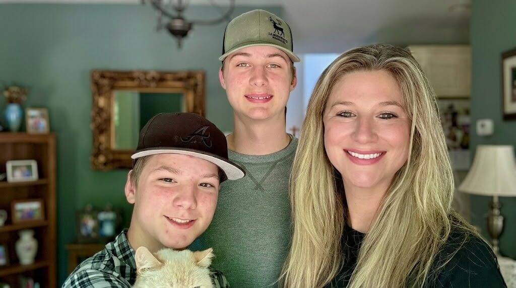 Three smiling people, two teenage boys wearing baseball caps and a blonde woman, with a cat in a home setting.
