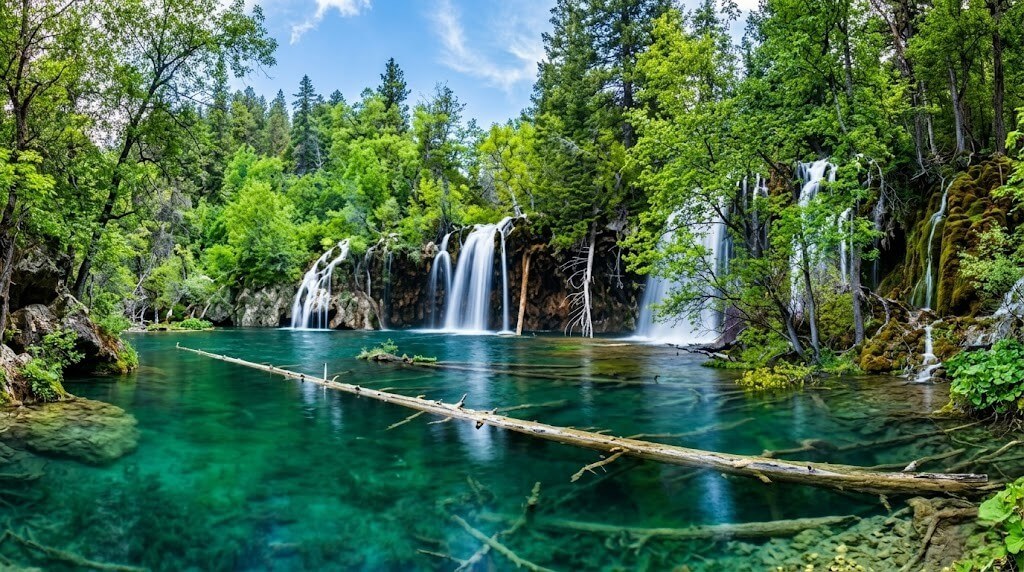 Clear turquoise lake with multiple waterfalls surrounded by lush green forest under a blue sky