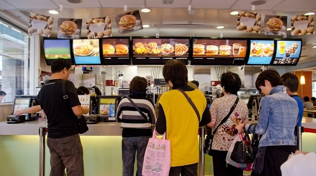 Customers standing in line ordering food at a fast-food restaurant counter with menu screens displaying burgers and desserts.