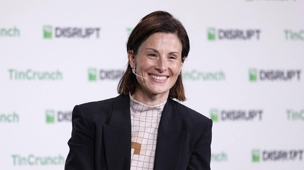 Woman smiling wearing a black blazer and checkered top speaking at TechCrunch Disrupt event