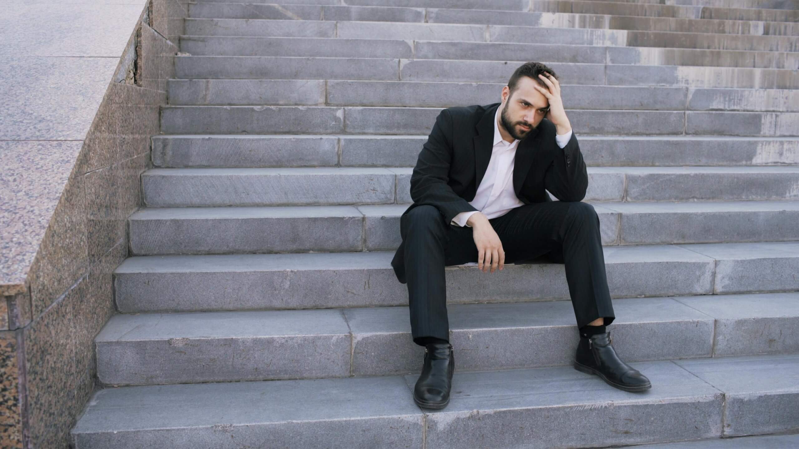 Man in black suit sitting on outdoor stone steps holding his head looking stressed